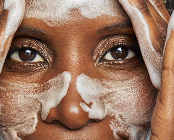 Detailný pohľad na zrelú ženu umývajúcu si rukami tvár, po celej tvári – na lícach, nose a čele – má bielu mydlovú penu.