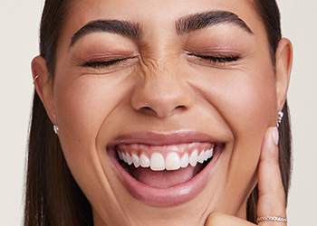 Mladá žena s výrazným obočím a dlhými rovnými hnedými vlasy, smeje sa, oči má zatvorené a nos pokrčený. 